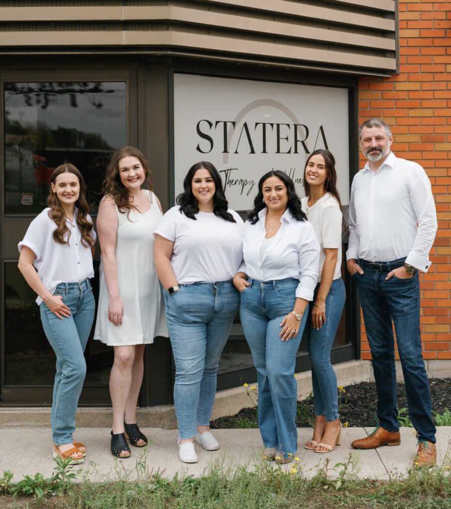 Group of six adults standing outside a clinic sign that reads STATERA Therapy and Wellness, wearing white tops and blue jeans.